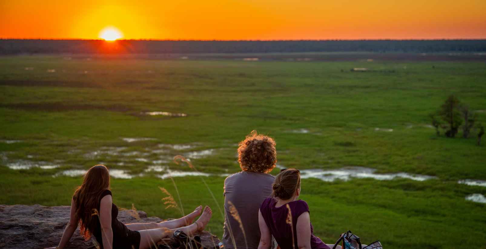 three people sitting on cliff overlooking wetlands as sun sets in distance